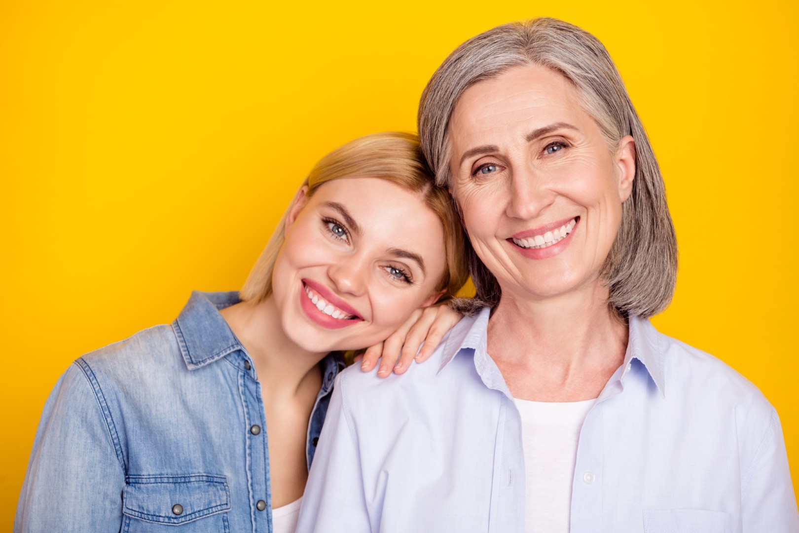 Photo portrait of daughter and mother smiling. They are standing against a vivid yellow backdrop.