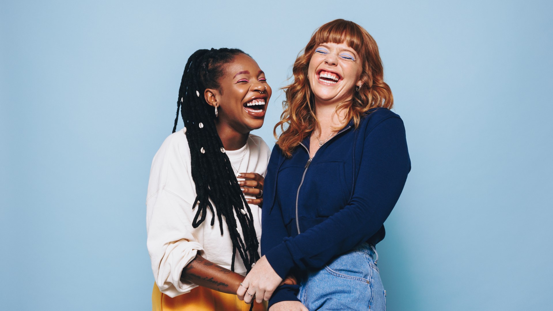 Best friends laughing cheerfully while standing together in a studio against a blue background.