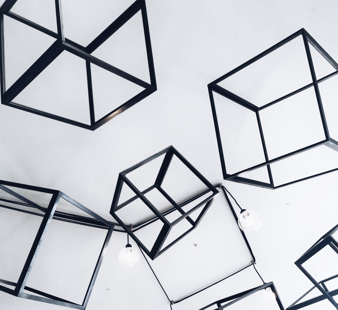 Des cadres cubiques en métal noir sont suspendus à un plafond blanc ; quelques ampoules apparentes sont également visibles parmi ces formes géométriques, créant ainsi un décor moderne et abstrait.