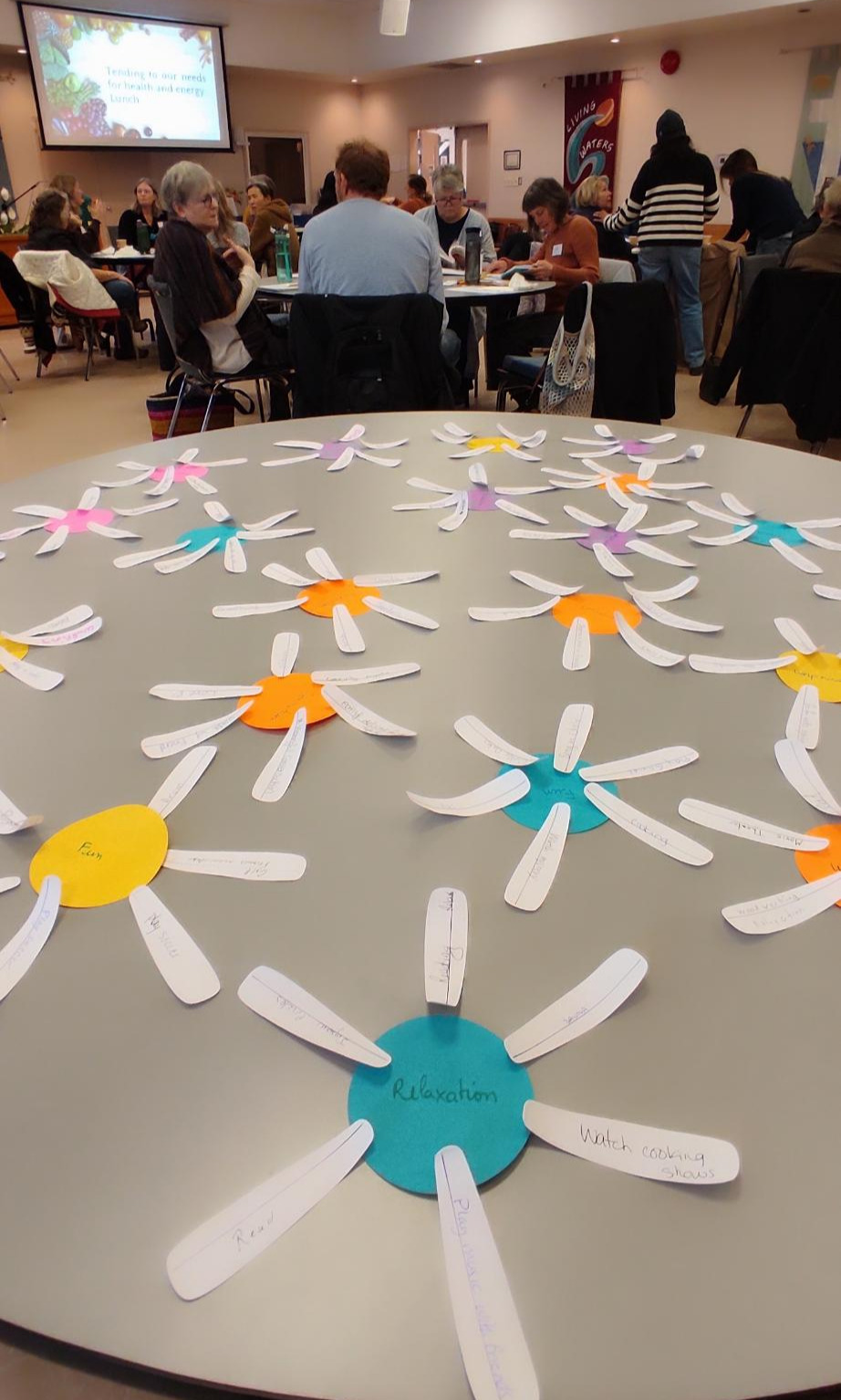People sit in groups at round tables in a large room. In the foreground, paper flowers with handwritten notes on each petal are spread out on a table.