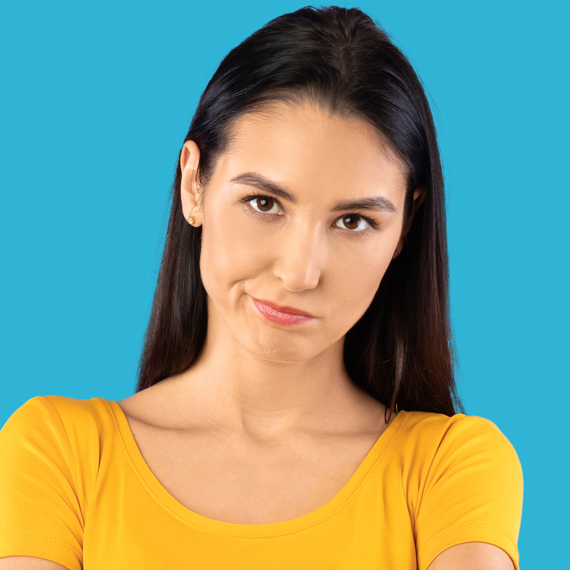 A woman with long dark hair wearing a yellow shirt stands against a blue background, looking forward with a slight frown and raised eyebrow