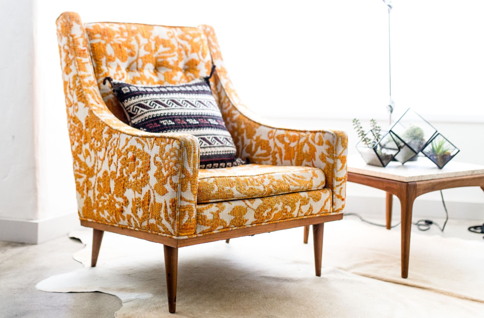 A vintage orange floral armchair with a patterned cushion sits next to a wooden side table holding geometric glass terrariums and small plants.